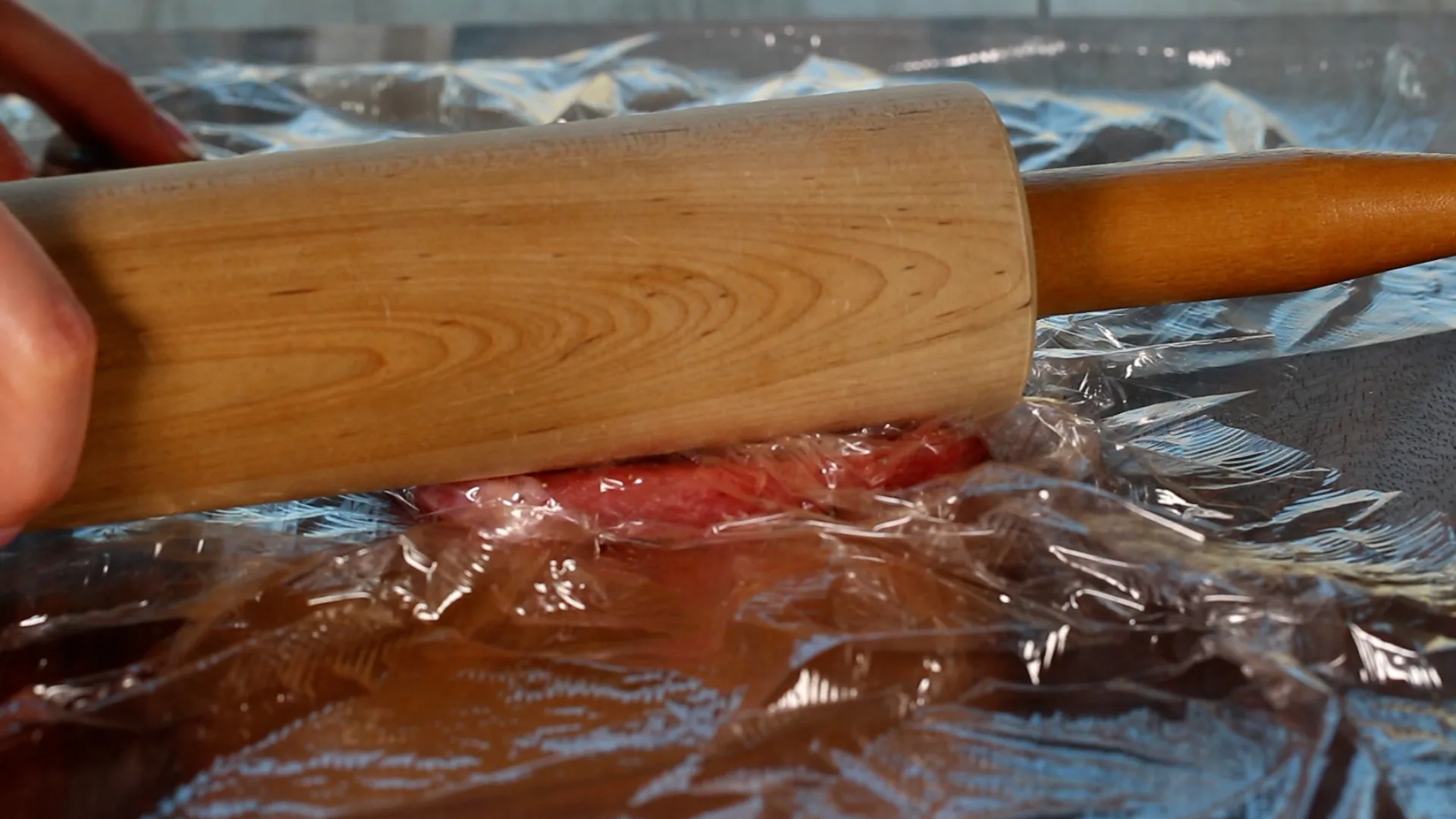 A rolling pin being used to tenderize the meat between plastic wrap.