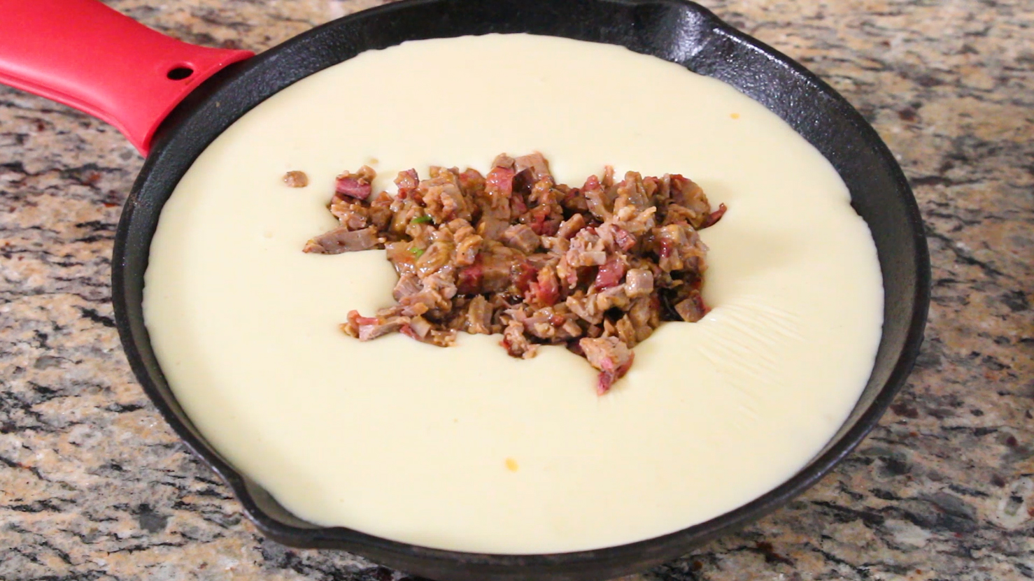 A cast iron skillet filled with queso blanco, being topped with chopped brisket morsels