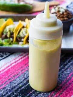 A squeeze bottle of creamy jalapeno sauce standing in front of a plate of tacos.