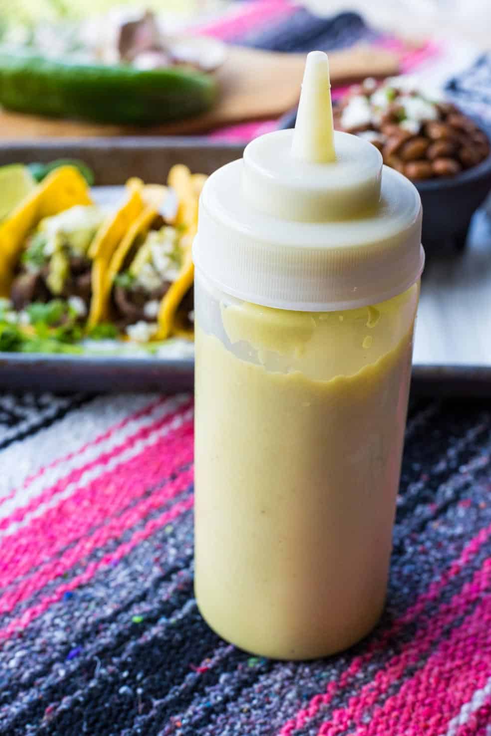 A squeeze bottle of creamy jalapeno sauce standing in front of a plate of tacos. 