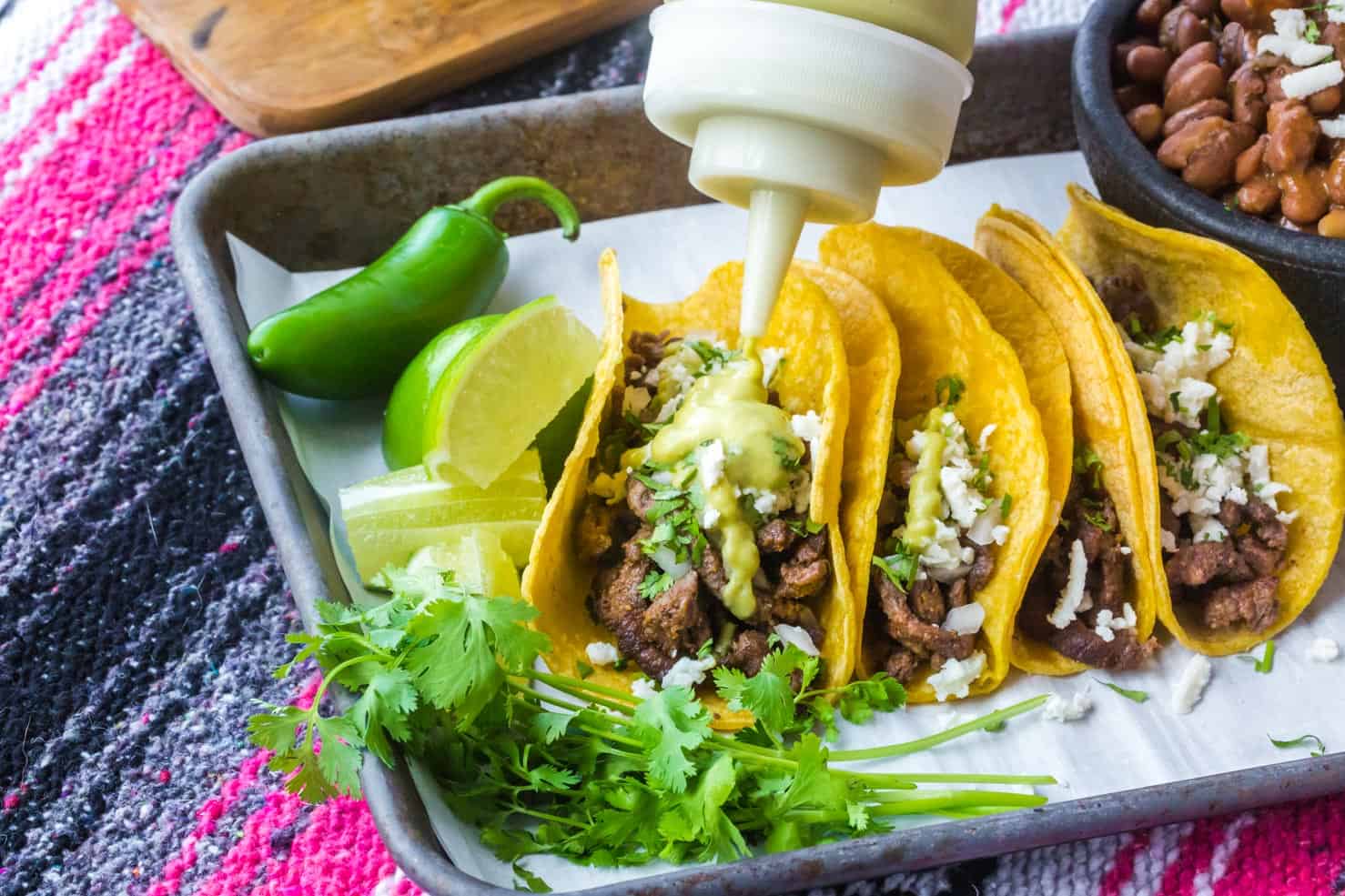 A platter of tacos on a pink serape table cloth. Creamy Jalapeno Sauce is being squeezed onto the tacos.