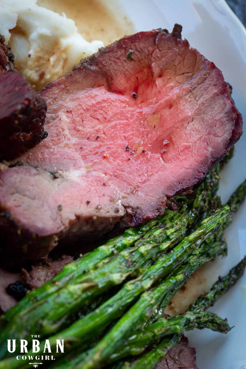 A close up of a white plate filled with mashed potatoes, sliced of pink and juicy smoked beef tenderloin medallions, and roasted asparagus. 