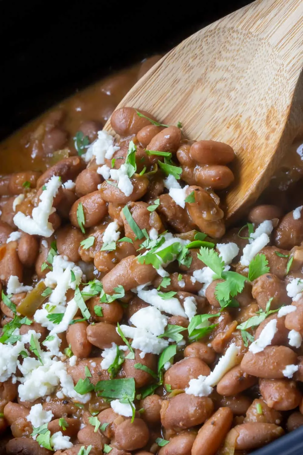 A close up of a crockpot full of glistening cooked pinto beans topped with cilantro and crema being served from a wooden spoon.