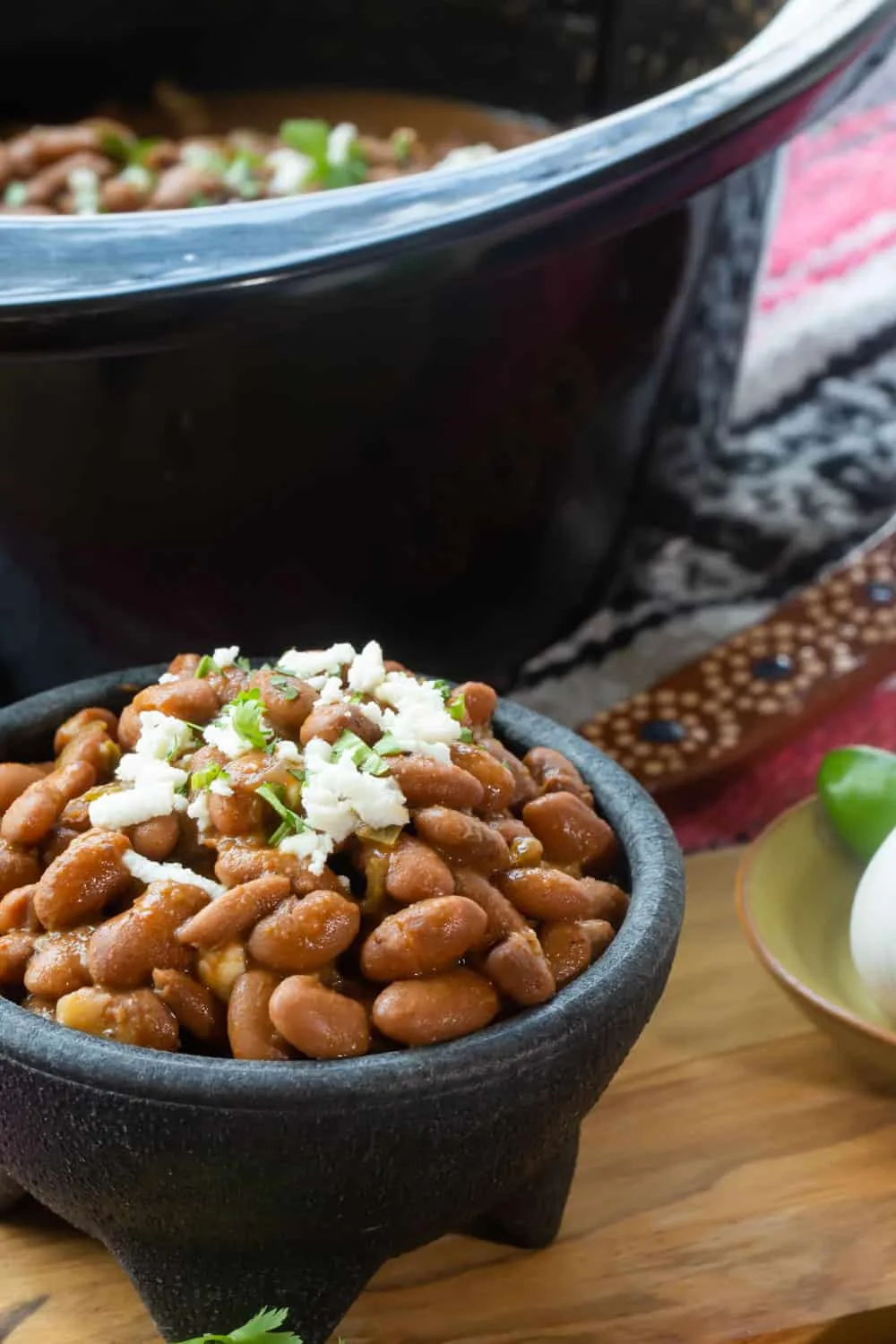A close up of a cup of beans, topped with garnishes, served in front of a crockpot. 