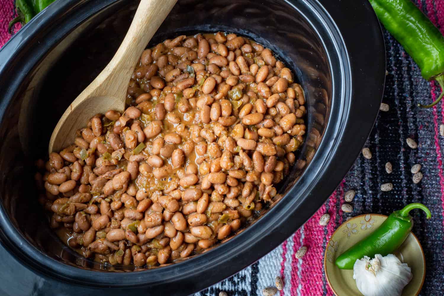 A crockpot full of glistening cooked pinto beans just cooked, served atop of festive mexican blanket.