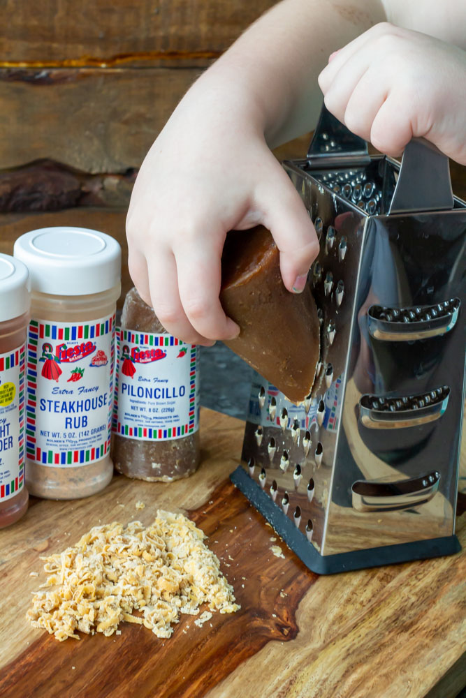 A child's hands grating fresh piloncillo brown sugar from the cone on a kitchen grater, an important step in preparing the pork rub.