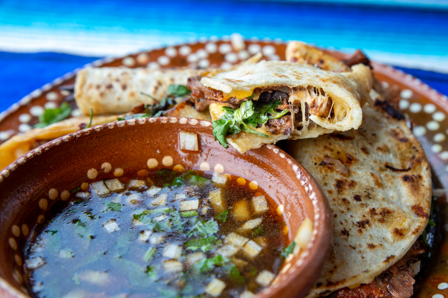 Half a birria taco open with the cheese oozing out, atop the birria sauce bowl.