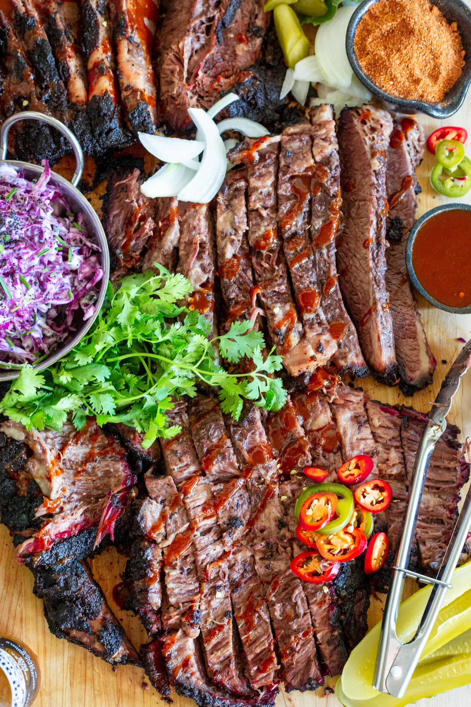 A platter of sliced brisket with coleslaw, spicy peppers, and serving utensils.