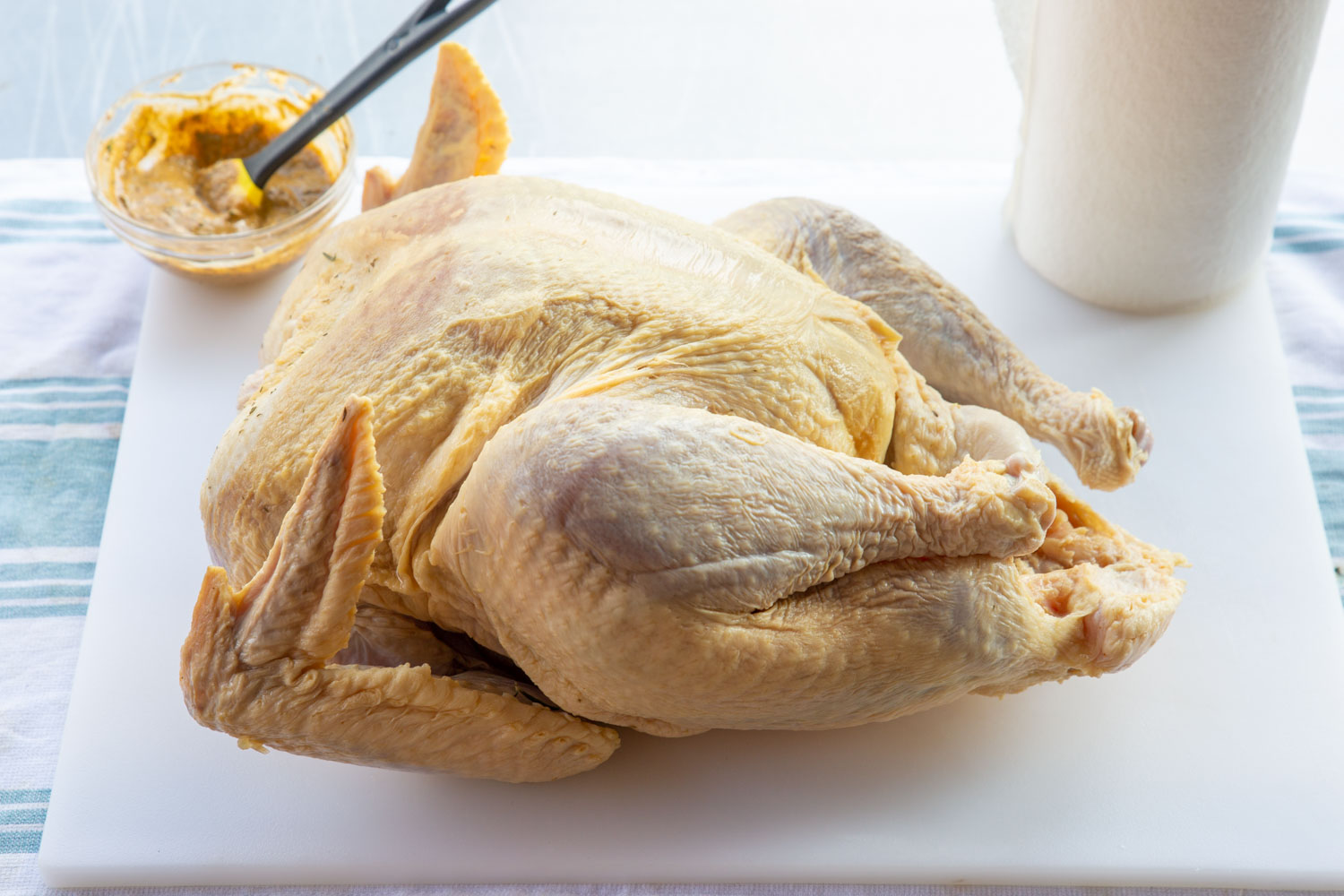 A raw turkey on a cutting board with a bowl of turkey paint behind it.