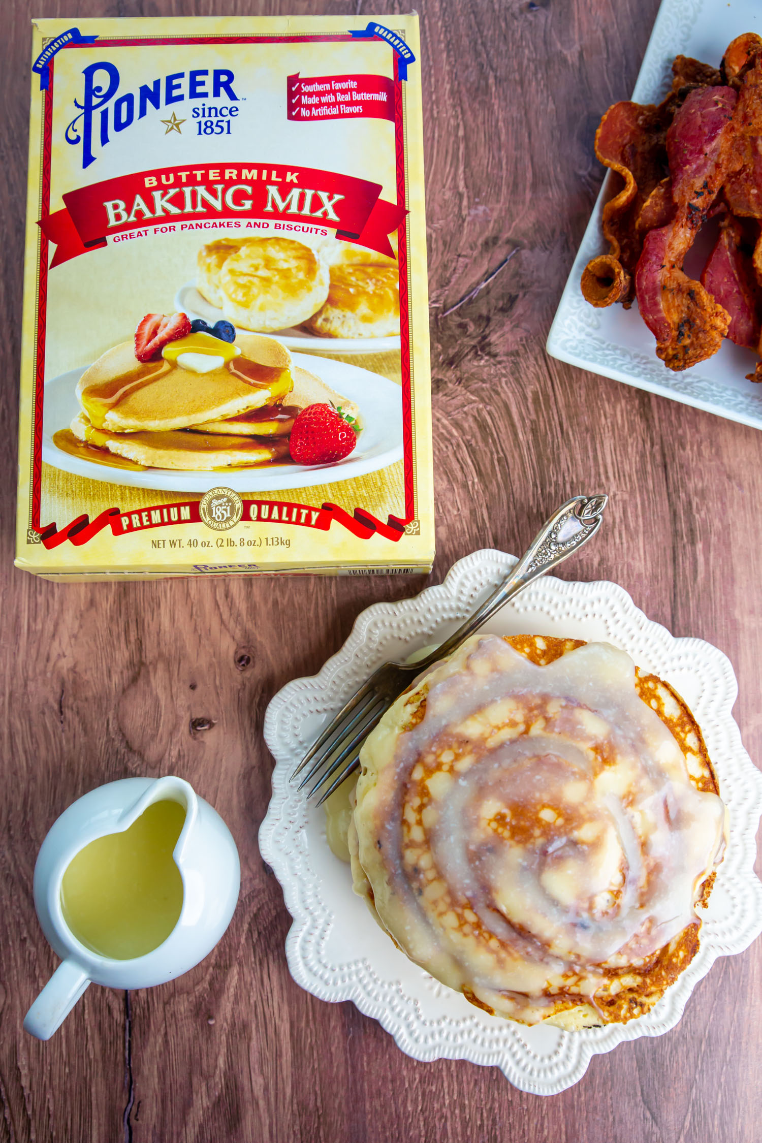 A box of pancake mix, a stack of pancakes, a cup of glaze and a plate of bacon on a wooden table.