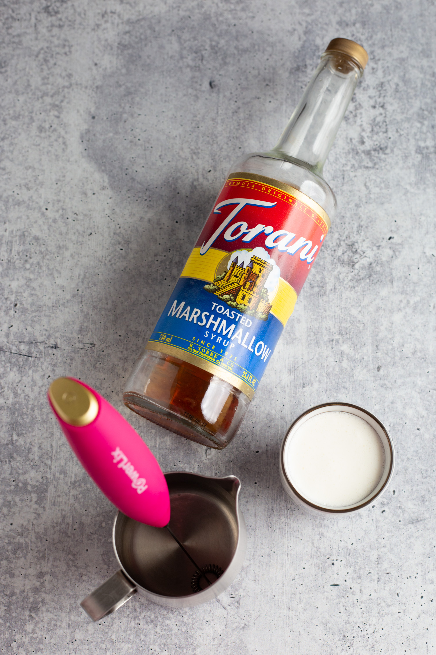 Torani Syrup, a cup of cream, and milk frothing pitcher, and a pink milk frother on a grey background