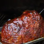 A smoked ham on a pellet smoker with bright red sauce and rub all over it.