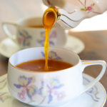 A tea cup being poured into by a tea kettle with a bright orange spiced tea drink.
