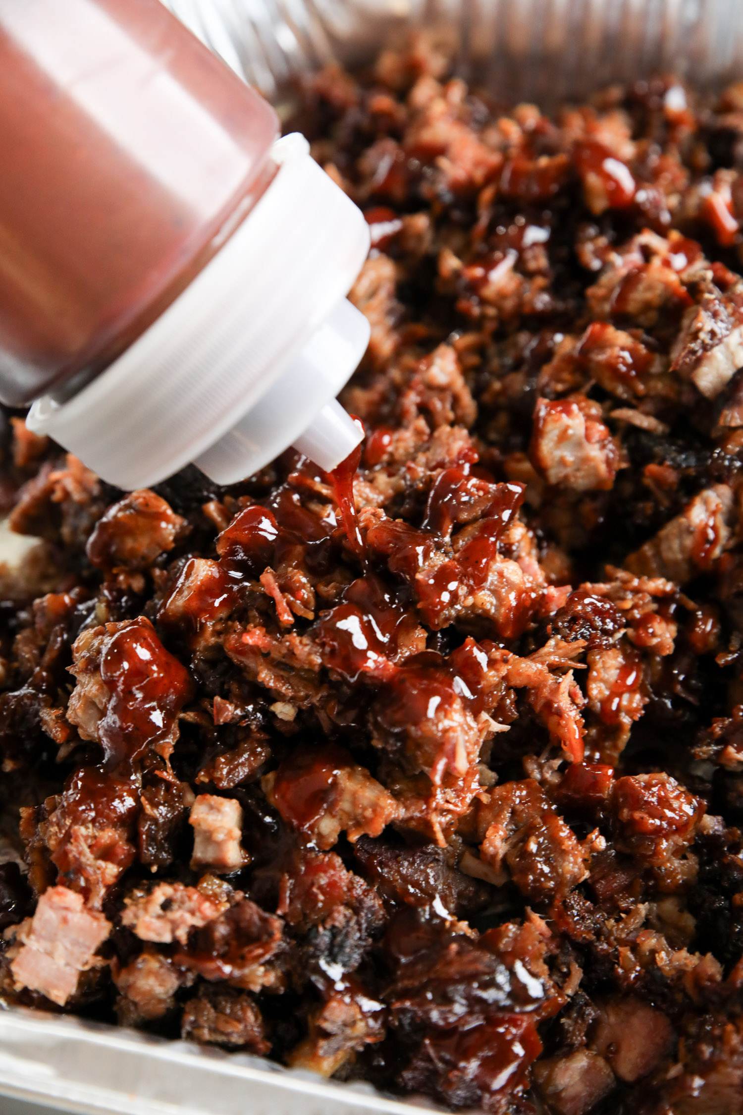An aluminum pan of chopped brisket being bound with a drizzle of barbecue sauce