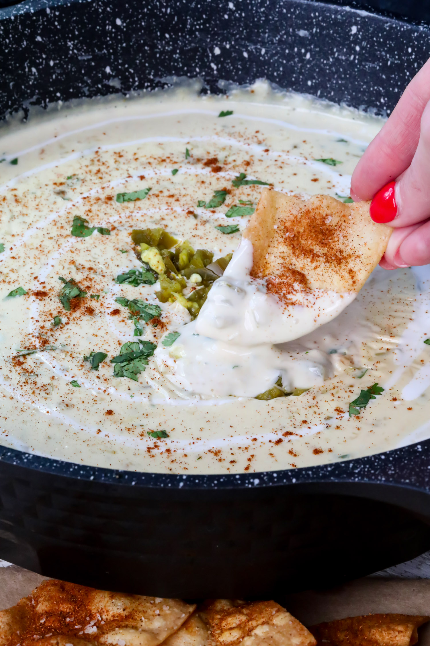 a hand dipping a tortilla chip into a bowl of white queso dip, with green chiles and seasonings on top