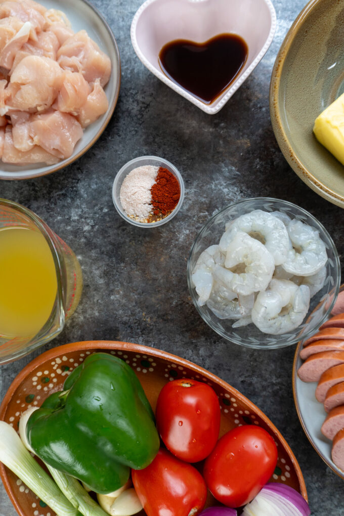 Pastalaya Ingedients: Raw Shrimp, Bell Peppers, Tomatoes, and Red Onion, Chicken Broth, Raw chicken, sausage, cajun spices all showcased in bowls.