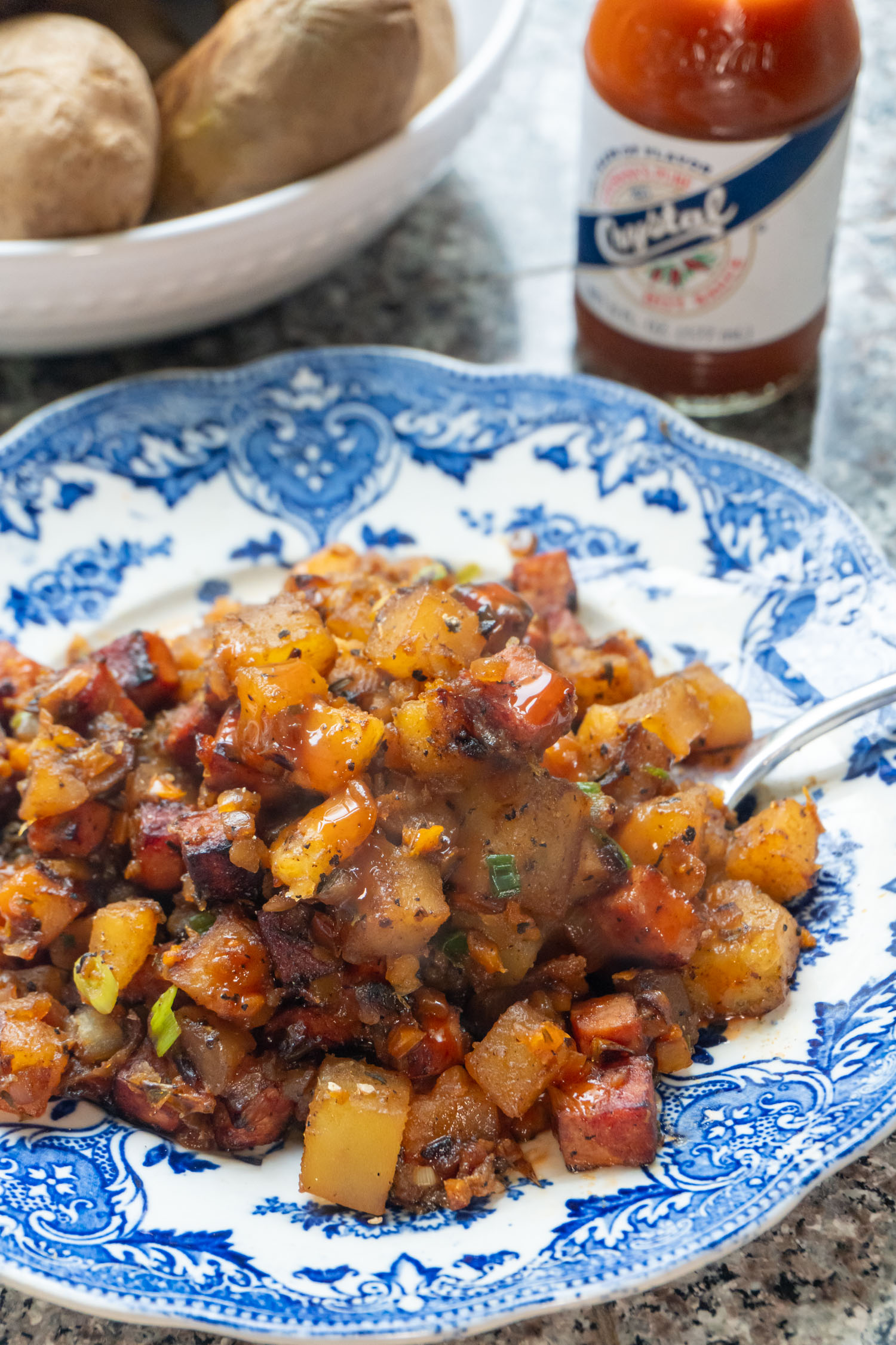 A blue and white floral plate filled with fried potatoes with crisp ham, topped with hot sauce