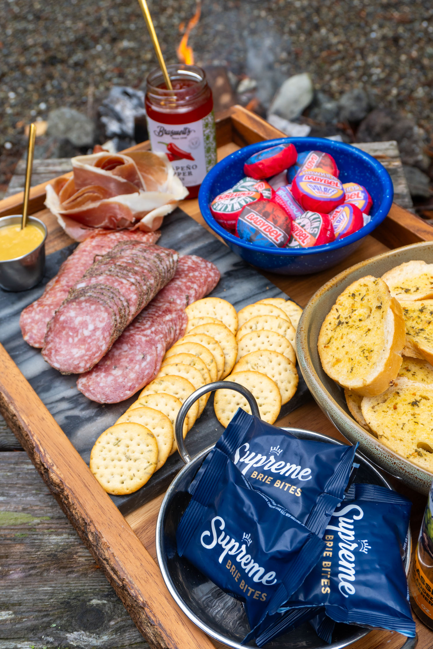 A large wooden platter of ingredients for savory smores displayed like a charcuterie board. Crackers, cheese, hot pepper jam, honeu mustard, salami, bread.