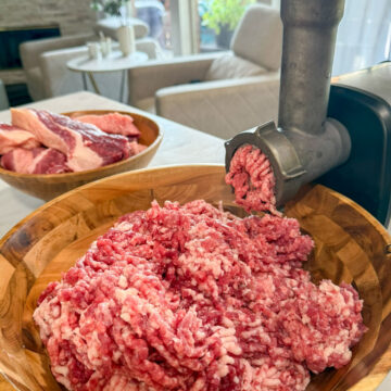 A meat grinder griding fresh brisket into a large wooden bowl in a modern kitchen.