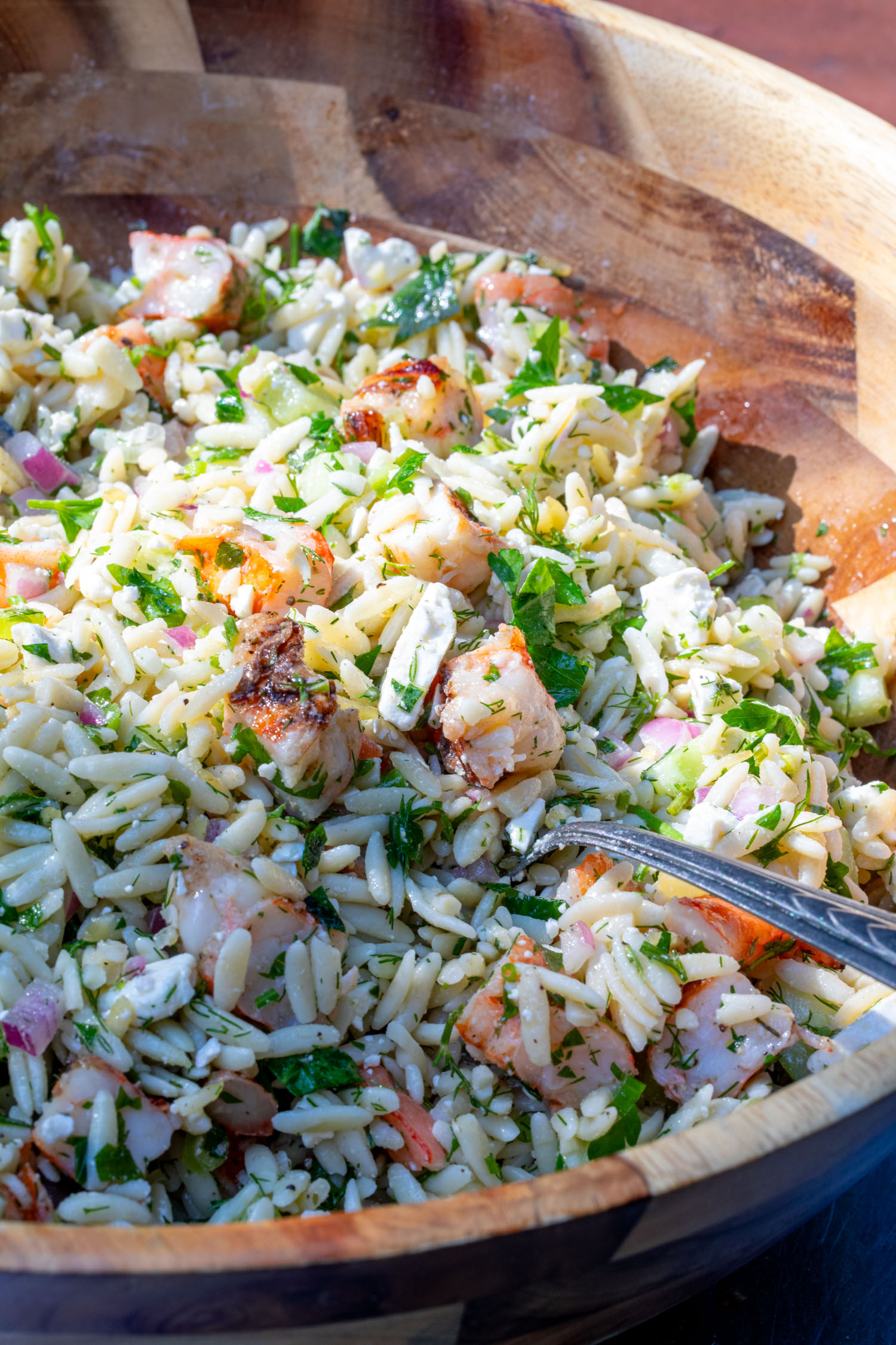 A wooden bowl of grilled shrimp orzo salad with fresh green herbs and feta cheese.